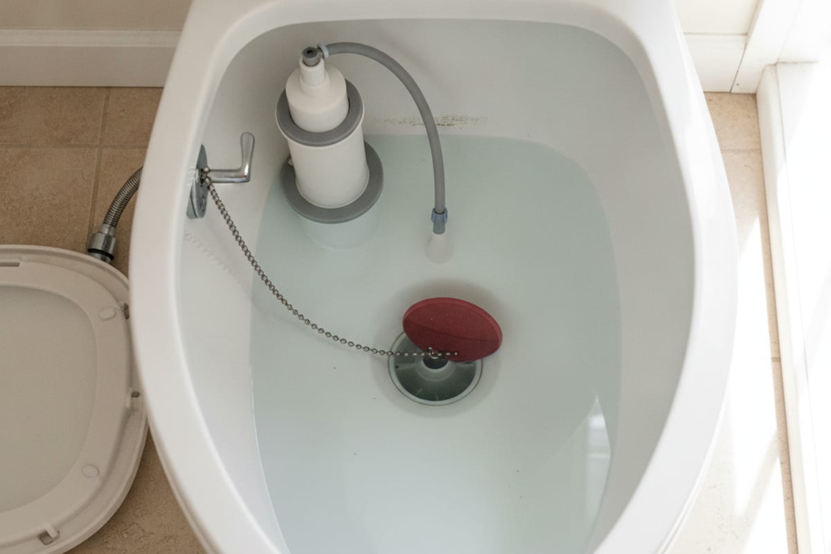 Open toilet tank in a Tampa Bay bathroom with the porcelain lid set aside on beige tile, showing the red rubber flapper on its seat, the lift chain running to the trip lever, and the white fill valve, float collar and overflow tube